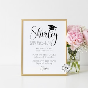 Op de afbeelding: Een ingelijst afstudeerbord met een gouden rand. Het bord leest "Shirley, She Can't Be Graduating" met cocktailrecepten. Een afstudeerhoed-afbeelding staat boven de naam. Een boeket roze bloemen staat rechts.