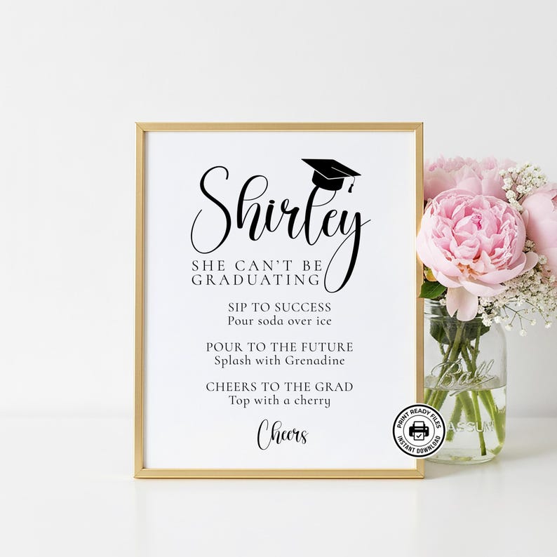 Op de afbeelding: Een ingelijst afstudeerbord met een gouden rand. Het bord leest "Shirley, She Can't Be Graduating" met cocktailrecepten. Een afstudeerhoed-afbeelding staat boven de naam. Een boeket roze bloemen staat rechts.