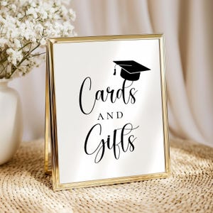 Op de afbeelding: Een goudkleurig bord met de woorden "Kaarten en Cadeaus" in elegant schrift, bekroond met een zwarte afstudeerpet illustratie. Het bord wordt weergegeven op een geweven oppervlak, met een vaas met witte bloemen op de achtergrond.