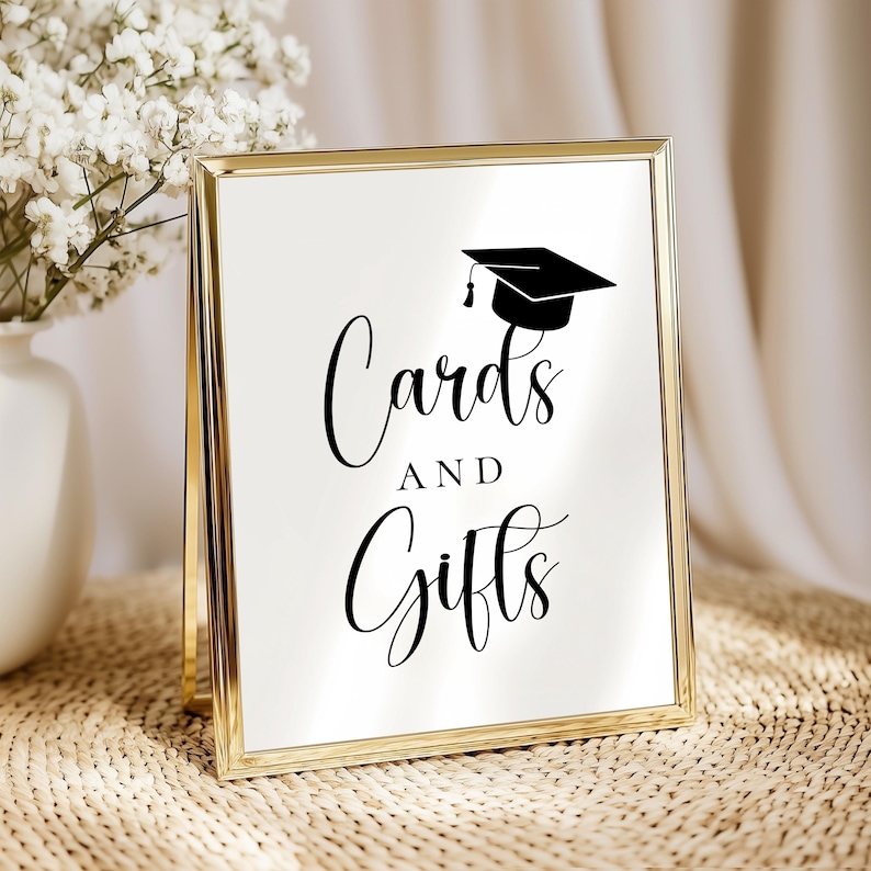 Op de afbeelding: Een goudkleurig bord met de woorden "Kaarten en Cadeaus" in elegant schrift, bekroond met een zwarte afstudeerpet illustratie. Het bord wordt weergegeven op een geweven oppervlak, met een vaas met witte bloemen op de achtergrond.