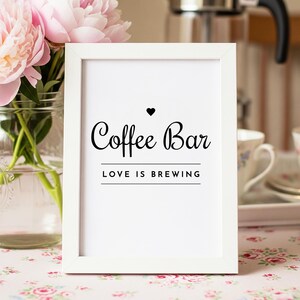 May include: A white framed sign with the words "Coffee Bar" in elegant black script, topped with a heart symbol. Below, the phrase "LOVE IS BREWING" is centered. Pink peonies in a glass vase are in the background.