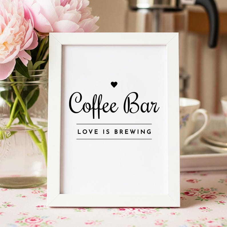 May include: A white framed sign with the words "Coffee Bar" in elegant black script, topped with a heart symbol. Below, the phrase "LOVE IS BREWING" is centered. Pink peonies in a glass vase are in the background.