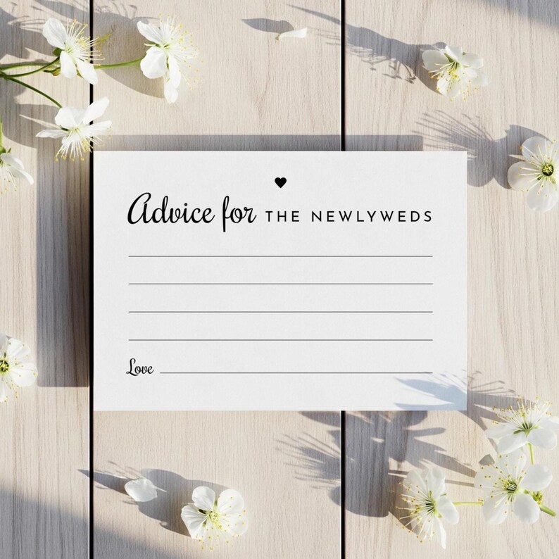May include: A white card with the text "Advice for the Newlyweds" in black script, with a small heart symbol. The card has lines for writing and the word "Love" at the bottom. White flowers are scattered around the card.