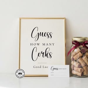May include: A gold-framed sign with cream background and black text reading Guess HOW MANY Corks Good Luck, accompanied by a small matching guess card and a clear glass jar filled with natural brown wine corks tied with a dark red ribbon.