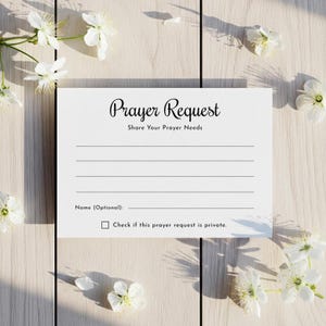 May include: A white card with the words "Prayer Request" in elegant script. The card includes lines for writing, the text "Share Your Prayer Needs," and a checkbox for privacy. White flowers and greenery surround the card on a light wood surface.