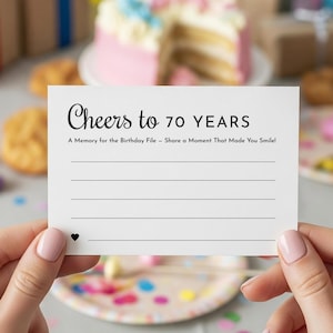 Può includere: Una carta bianca con la scritta "Cheers to 70 YEARS" in corsivo nero. Sotto, la carta recita "A Memory for the Birthday File - Share a Moment That Made You Smile!" e ha delle righe per scrivere. Un piccolo cuore nero è in basso.
