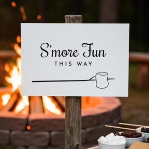 May include: A white sign on a wooden post with the text "S'more Fun THIS WAY" in black script. A line leads to a marshmallow illustration. A campfire burns brightly in the background, creating a warm ambiance.