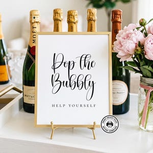 May include: A gold-framed sign with the words "Pop the Bubbly" in elegant script, accompanied by "Help Yourself." The sign is displayed on a gold stand, with champagne bottles and pink flowers in the background. A print-ready files logo is in the bottom right.