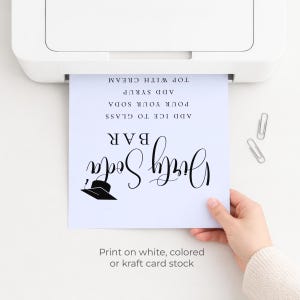 Op de afbeelding: Een wit bedrukt bord met de woorden "Dirty Soda Bar" in elegant zwart schrift. Instructies omvatten het toevoegen van ijs, frisdrank, siroop en room. Het bord wordt door een hand bij een printer gehouden.