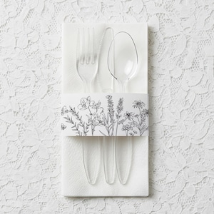 Könnte beinhalten: Klares Kunststoff-Besteck-Set mit Gabel, Messer und Löffel auf einer weißen Serviette, zusammengehalten von einem Papierband mit einem schwarz-weißen Blumendesign. Ideal für Picknicks.