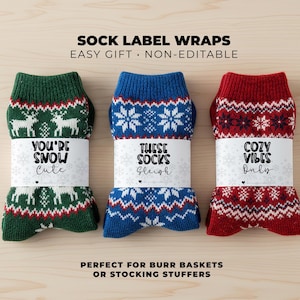 Pode incluir: Três pares de meias de Natal dobradas com etiquetas decorativas. Um par é verde com renas brancas, um é azul com flocos de neve e um é vermelho com estrelas. Os rótulos dizem "You're Snow Cute", "These Socks Sleigh" e "Cozy Vibes Only".