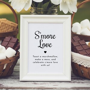 May include: A white framed sign with the text "S'more Love" and a heart symbol. The sign includes the text "Toast a marshmallow, make a mess, and celebrate s'more love with us!" Baskets of marshmallows, chocolate, and graham crackers are in the background.