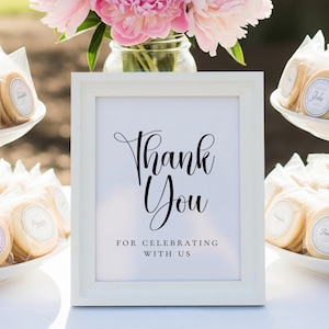 Puede incluir: Un letrero enmarcado en blanco con las palabras "Thank You" en elegante escritura negra, con la frase "For Celebrating With Us" debajo. Flores rosas y galletas envueltas en el fondo sugieren una celebraci&oacute;n.