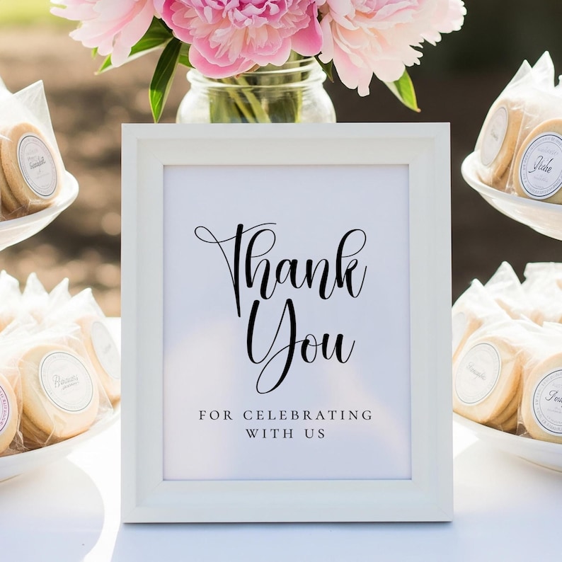 Puede incluir: Un letrero enmarcado en blanco con las palabras "Thank You" en elegante escritura negra, con la frase "For Celebrating With Us" debajo. Flores rosas y galletas envueltas en el fondo sugieren una celebraci&oacute;n.