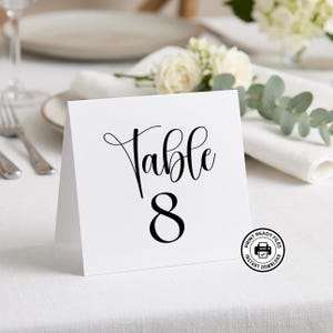 Può includere: Un biglietto bianco con il numero del tavolo con la parola "Table" in elegante corsivo e il numero "8" in grassetto. Il biglietto è esposto su una tovaglia bianca, con una composizione floreale sullo sfondo.