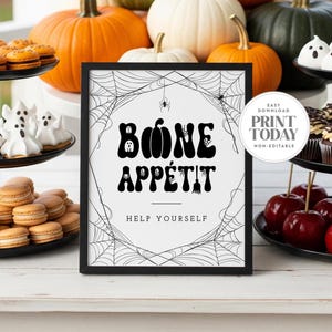 May include: A framed Halloween sign with the words "Bone Appétit" in black, surrounded by a spiderweb design. The sign is on a white surface, with pumpkins and treats in the background. The sign also says "Help Yourself".
