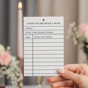 Puede incluir: Una tarjeta blanca con el texto "ADVICE FOR THE BRIDE & GROOM" y el título "A New Chapter Begins". La tarjeta tiene líneas para escribir consejos y deseos. Un símbolo de corazón está encima del texto. La tarjeta es sostenida por una persona.