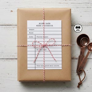 Könnte beinhalten: Ein in braunes Papier verpacktes Paket, das mit rot-weißem Garn verschnürt ist. Eine weiße Karte mit dem Text "BLIND DATE WITH A COOKBOOK" ist angebracht. Kupfer-Messlöffel befinden sich auf der rechten Seite. Die Karte enthält Aufforderungen für Genre, Stimmung, Datum und Geschmacksrichtungen.