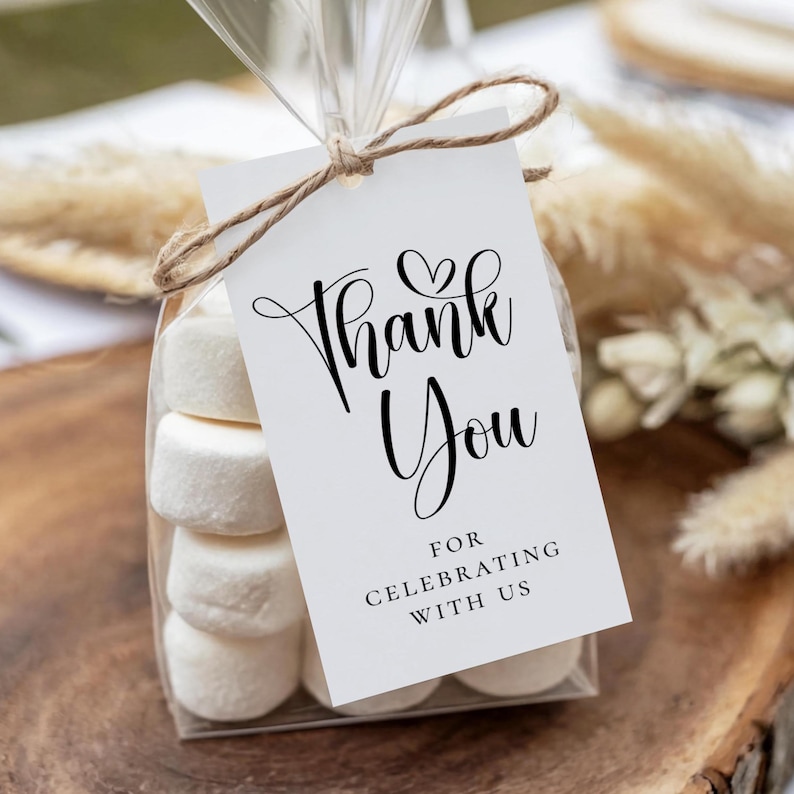 Puede incluir: Una bolsa transparente llena de malvaviscos blancos, atada con hilo. Una etiqueta blanca con la inscripci&oacute;n en negro "Thank You" y "For Celebrating With Us". El fondo es una superficie de madera.