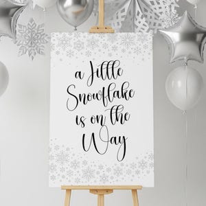 May include: A white sign on a wooden easel with the text "a little Snowflake is on the Way" in black script. The sign is decorated with snowflake designs. Silver balloons and snowflake decorations are in the background.