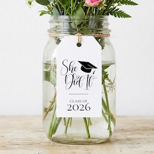 Op de afbeelding: Een glazen pot gevuld met water en verse bloemen, waaronder roze rozen en witte madeliefjes. Een wit label met de tekst "She Did It" en een afstudeerhoed-afbeelding is om de pot gebonden, met de woorden "Class of 2026" eronder.