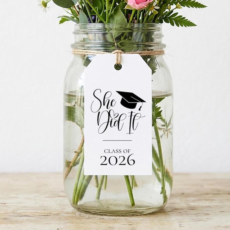 Op de afbeelding: Een glazen pot gevuld met water en verse bloemen, waaronder roze rozen en witte madeliefjes. Een wit label met de tekst "She Did It" en een afstudeerhoed-afbeelding is om de pot gebonden, met de woorden "Class of 2026" eronder.