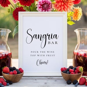Puede incluir: Un letrero enmarcado en blanco con las palabras "Sangria Bar" y las instrucciones "Vierta el vino, cubra con fruta, &iexcl;Salud!" se muestra. Dos jarras de vidrio de sangr&iacute;a y cuencos de madera con fruta est&aacute;n en primer plano. Flores coloridas est&aacute;n en el fondo.