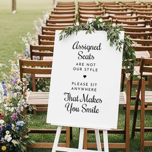 May include: A white sign on an easel with the text "Assigned Seats Are Not Our Style. Please Sit Anywhere That Makes You Smile." The sign is decorated with greenery and is set up in front of rows of wooden chairs at an outdoor event.