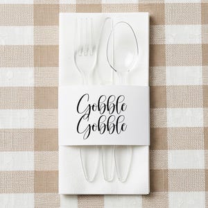 May include: A clear plastic fork, knife, and spoon set on a white napkin, held together by a white band with the words "Gobble Gobble" in black script. The background is a tan and white checkered pattern.