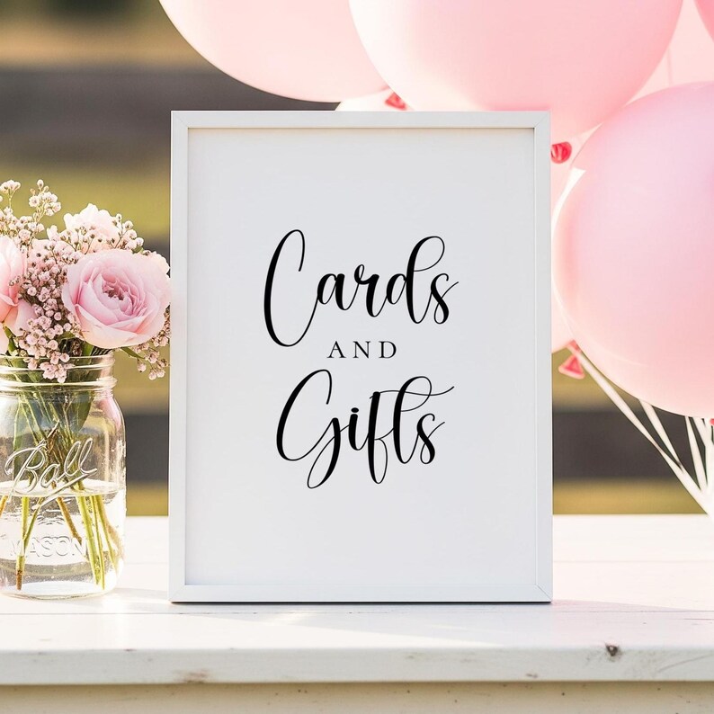 Puede incluir: Un letrero enmarcado blanco con las palabras "Cards and Gifts" en elegante escritura negra. El letrero est&aacute; sobre una superficie blanca, con globos rosas y un tarro de cristal con flores rosas en el fondo.
