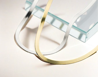 May include: Two snake chains, one gold and one silver, are displayed on a glass surface. The chains are both flat and have a slightly textured surface.