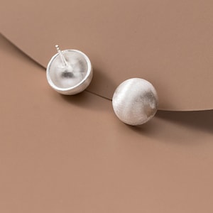 Peut inclure: Une paire de boucles d'oreilles en argent avec une surface lisse et arrondie. Les boucles d'oreilles sont sur un fond brun.