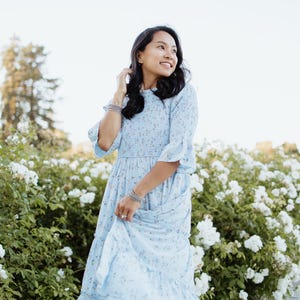 Puede incluir: Un vestido azul claro con estampado floral, mangas con volantes y una falda escalonada. El vestido lo lleva una persona de pie frente a un rosal blanco. El vestido tiene un corpi&ntilde;o ajustado y una falda fluida.