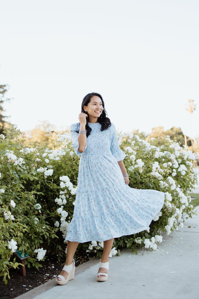Puede incluir: Vestido azul claro con estampado floral, falda escalonada y mangas cortas. El vestido se lleva con sandalias de cu&ntilde;a blancas. El fondo presenta flores blancas y vegetaci&oacute;n.