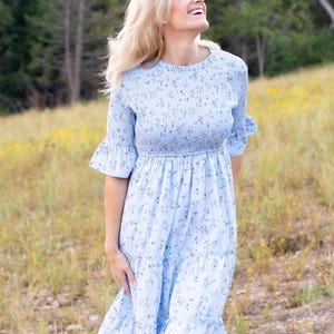 Puede incluir: Un vestido azul claro con estampado floral. El vestido tiene un corpi&ntilde;o fruncido, mangas cortas con pu&ntilde;os con volantes y una falda escalonada. El vestido es hasta la rodilla y parece estar hecho de una tela ligera.