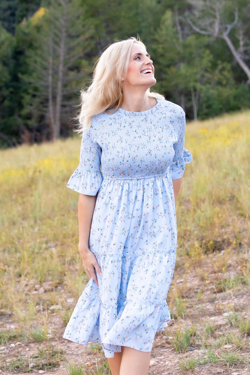 Puede incluir: Un vestido azul claro con estampado floral. El vestido tiene un corpi&ntilde;o fruncido, mangas cortas con pu&ntilde;os con volantes y una falda escalonada. El vestido es hasta la rodilla y parece estar hecho de una tela ligera.
