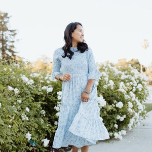 Puede incluir: Un vestido azul claro con estampado floral, falda escalonada y mangas con volantes. El vestido se lleva con sandalias blancas. El fondo presenta flores blancas y vegetaci&oacute;n, creando una est&eacute;tica suave y femenina.
