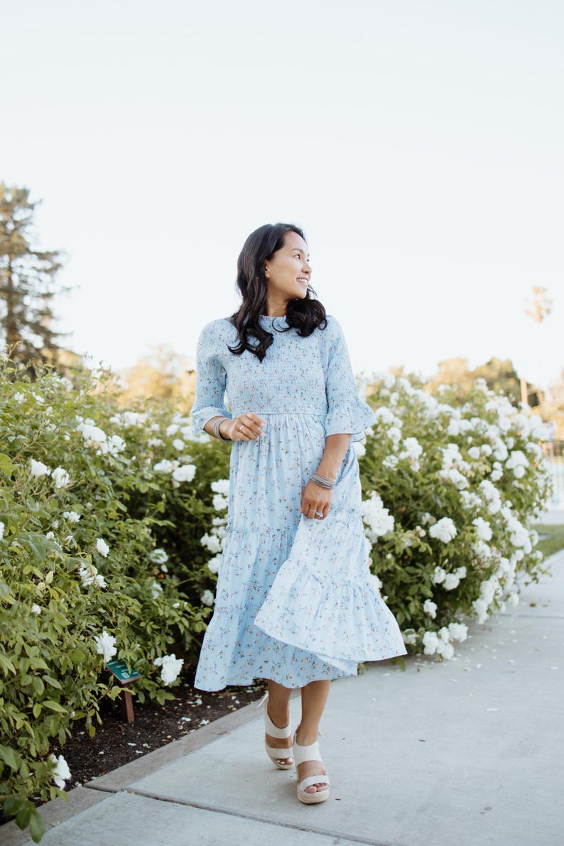 Puede incluir: Un vestido azul claro con estampado floral, falda escalonada y mangas con volantes. El vestido se lleva con sandalias blancas. El fondo presenta flores blancas y vegetaci&oacute;n, creando una est&eacute;tica suave y femenina.