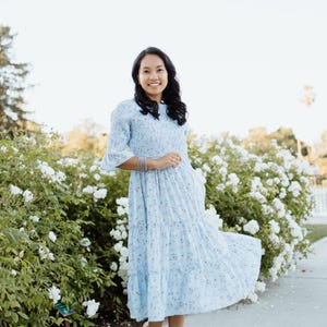 Puede incluir: Un vestido azul claro con estampado floral, falda escalonada y mangas cortas. El vestido se lleva con sandalias de plataforma blancas. El fondo presenta flores blancas y vegetaci&oacute;n, creando un ambiente suave y natural.