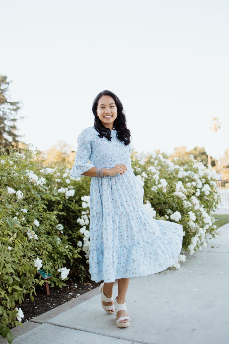 Puede incluir: Un vestido azul claro con estampado floral, falda escalonada y mangas cortas. El vestido se lleva con sandalias de plataforma blancas. El fondo presenta flores blancas y vegetaci&oacute;n, creando un ambiente suave y natural.