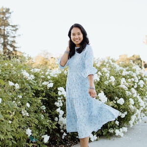 Puede incluir: Vestido midi azul claro con estampado floral y mangas con volantes. El vestido llega a la rodilla y tiene una falda escalonada. La persona lleva sandalias de plataforma blancas. El fondo presenta flores blancas y vegetaci&oacute;n.