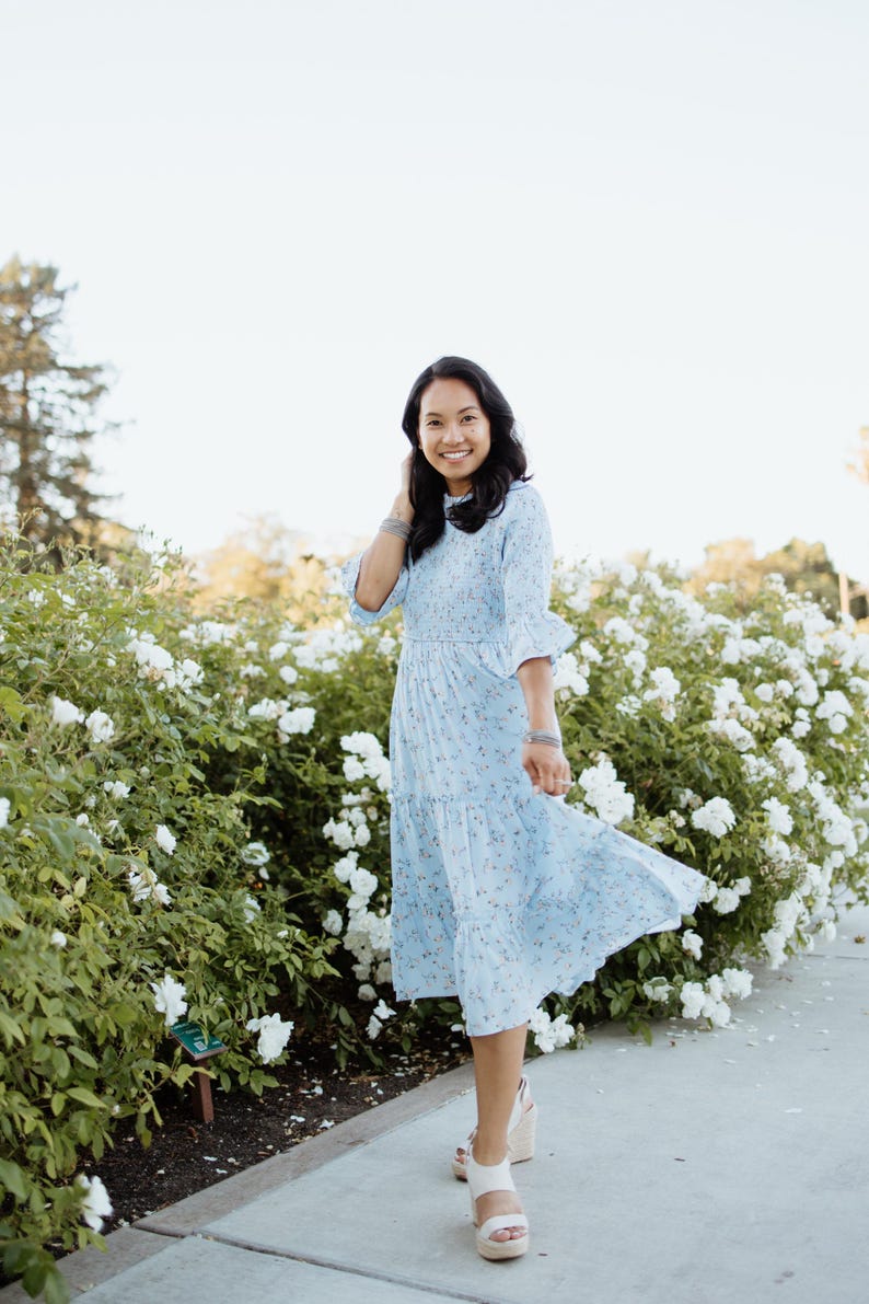 Puede incluir: Vestido midi azul claro con estampado floral y mangas con volantes. El vestido llega a la rodilla y tiene una falda escalonada. La persona lleva sandalias de plataforma blancas. El fondo presenta flores blancas y vegetaci&oacute;n.