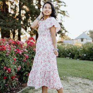 May include: A white floral print dress with pink flowers and ruffled sleeves. The dress is knee-length and paired with white wedge sandals. The background features a garden with red roses and green grass.