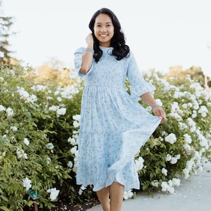 Puede incluir: Vestido azul claro con estampado floral, mangas de codo y falda escalonada. El vestido se lleva con sandalias de plataforma blancas. El fondo presenta un rosal blanco y una pasarela pavimentada.