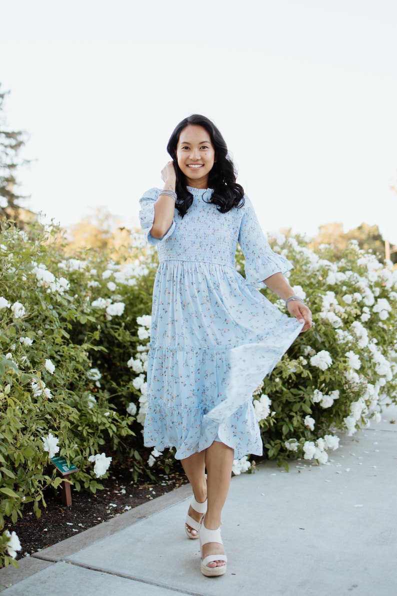 Puede incluir: Vestido azul claro con estampado floral, mangas de codo y falda escalonada. El vestido se lleva con sandalias de plataforma blancas. El fondo presenta un rosal blanco y una pasarela pavimentada.