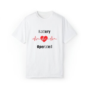 Op de afbeelding: Wit T-shirt met de tekst "Battery Operated" in zwarte letters. Een rood hart staat in het midden van een rode hartslaglijn. Het shirt heeft een ronde hals.