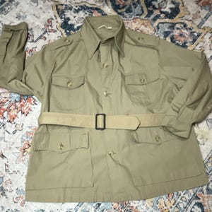 May include: A tan long-sleeve jacket featuring a pointed collar, shoulder epaulets, and a front button closure. The garment includes four front pockets with buttoned flaps and a matching tan belt with a rectangular black buckle at the waist.