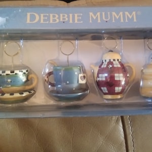 May include: Set of four ceramic teacup and teapot themed place card holders. The holders are in a blue and white checkered pattern with red accents. The set includes two teacups, a teapot, and a honey pot. The holders are packaged in a blue box with the words "Debbie Mumm" printed on the top.