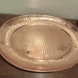 May include: Oval copper serving tray with hammered texture and two brass handles. The tray is a warm copper color and has a slightly raised rim. A decorative piece for serving food or displaying items.