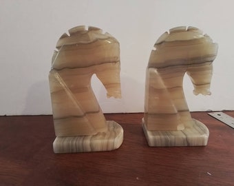 May include: A pair of bookends made of light brown and white striped alabaster, shaped like horse heads.